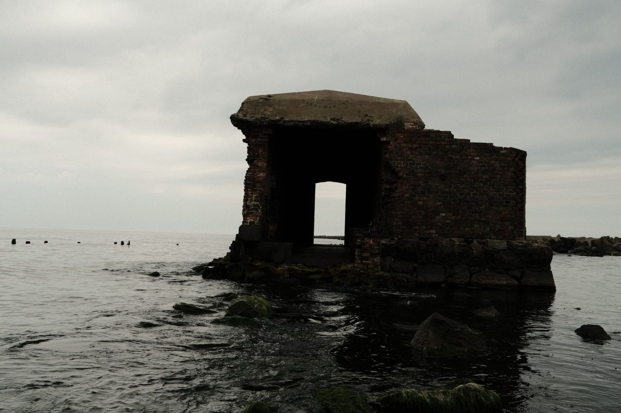 A roofless brick-and-concrete bunker with a doorway stands in shallow water under a grey sky.