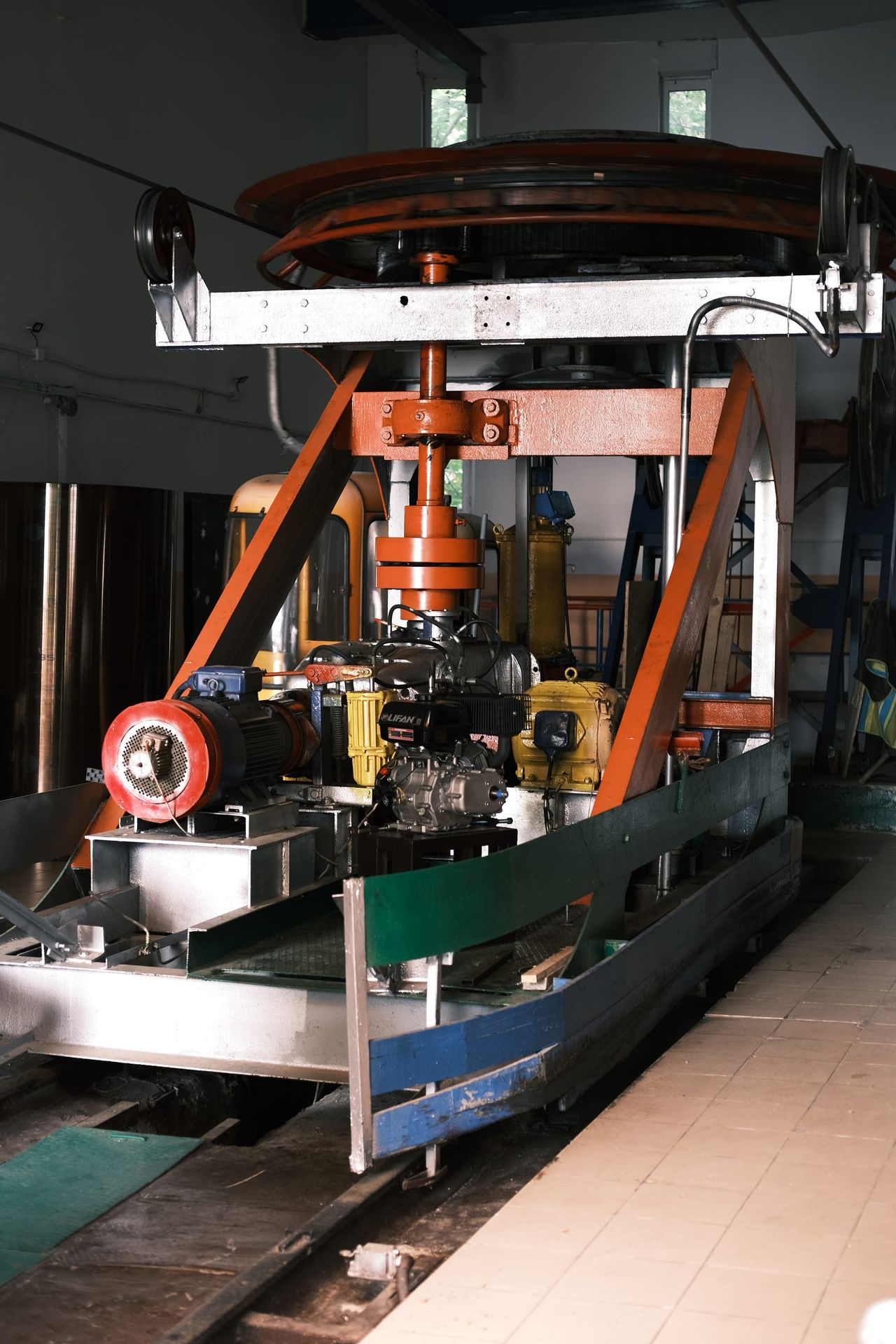 El mecanismo motor de un teleférico, con una gran rueda, un motor y un bastidor naranja, sobre raíles en una sala de estación con suelo de baldosas.