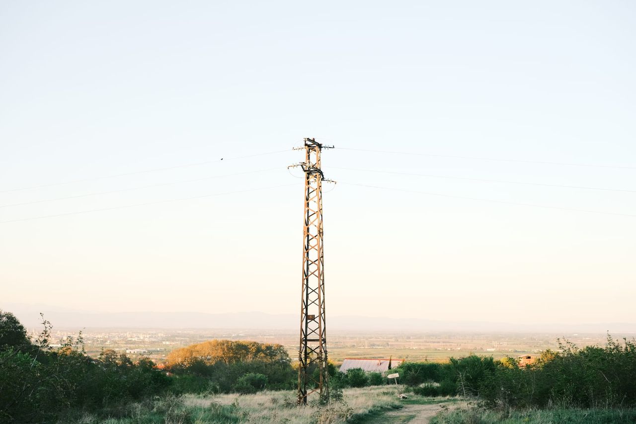 Slender lattice utility tower rises above trees and a distant skyline.