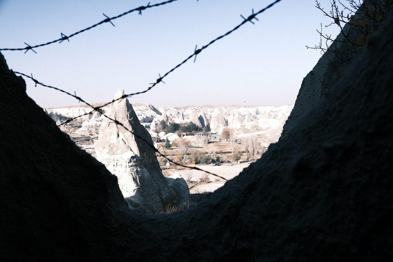 Distant buildings framed by rough dark structures and barbed wire.