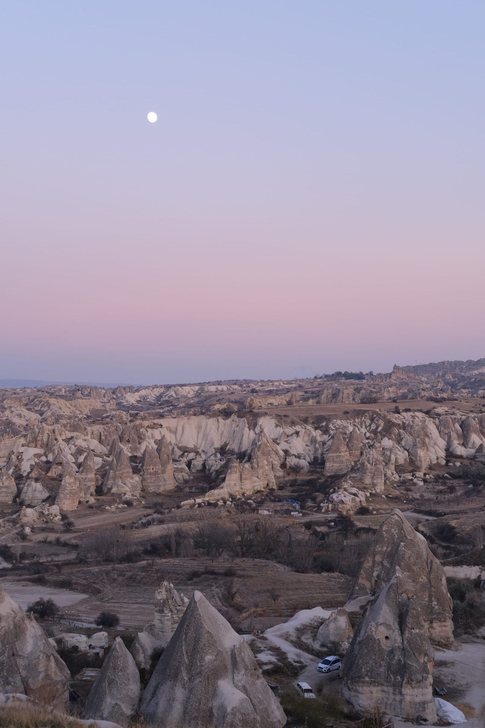 Moon above Cappadocia rock formations beneath a pale pink and blue twilight sky