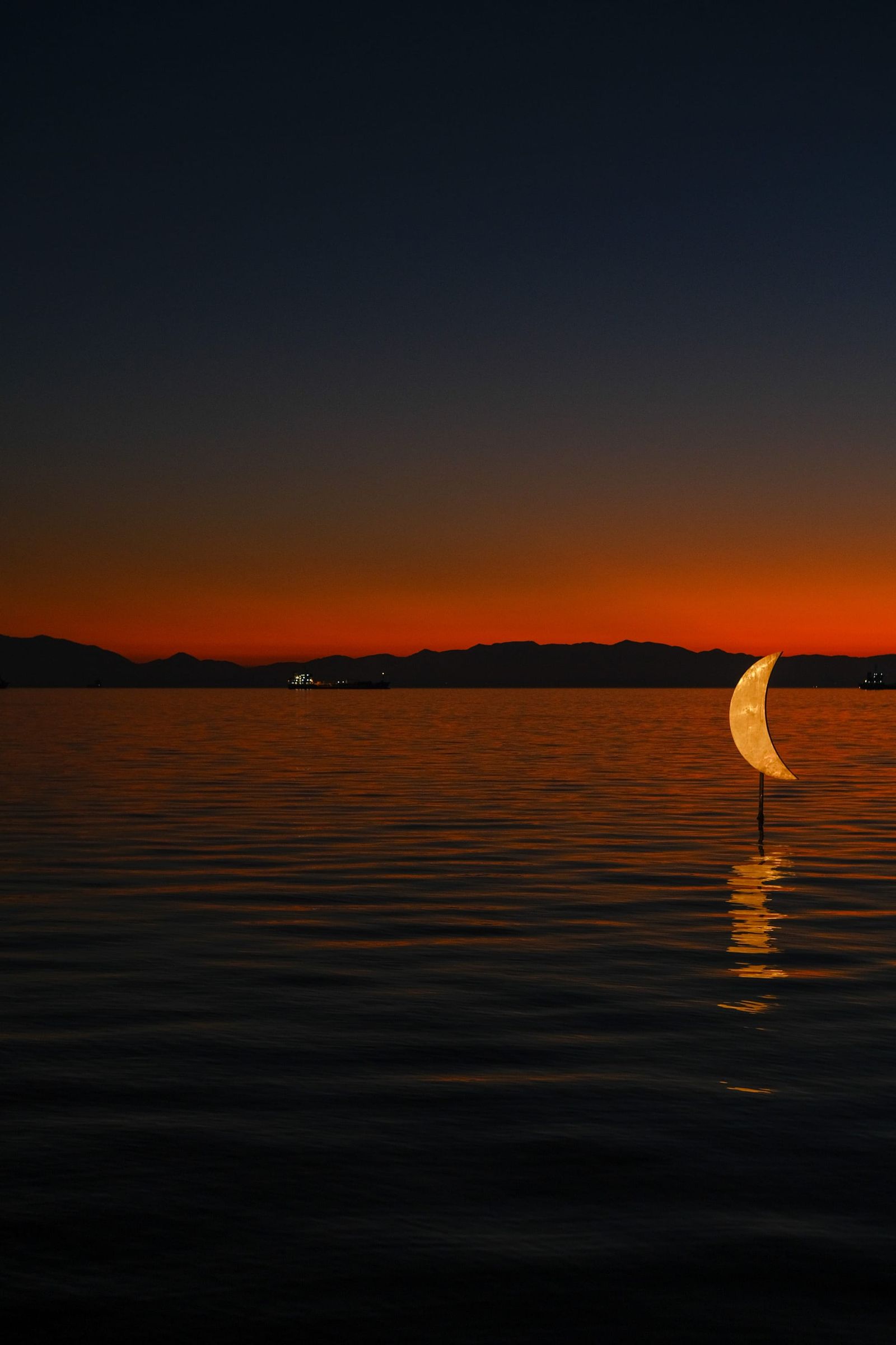 Crescent-shaped light reflected on calm water beneath a deep orange twilight sky