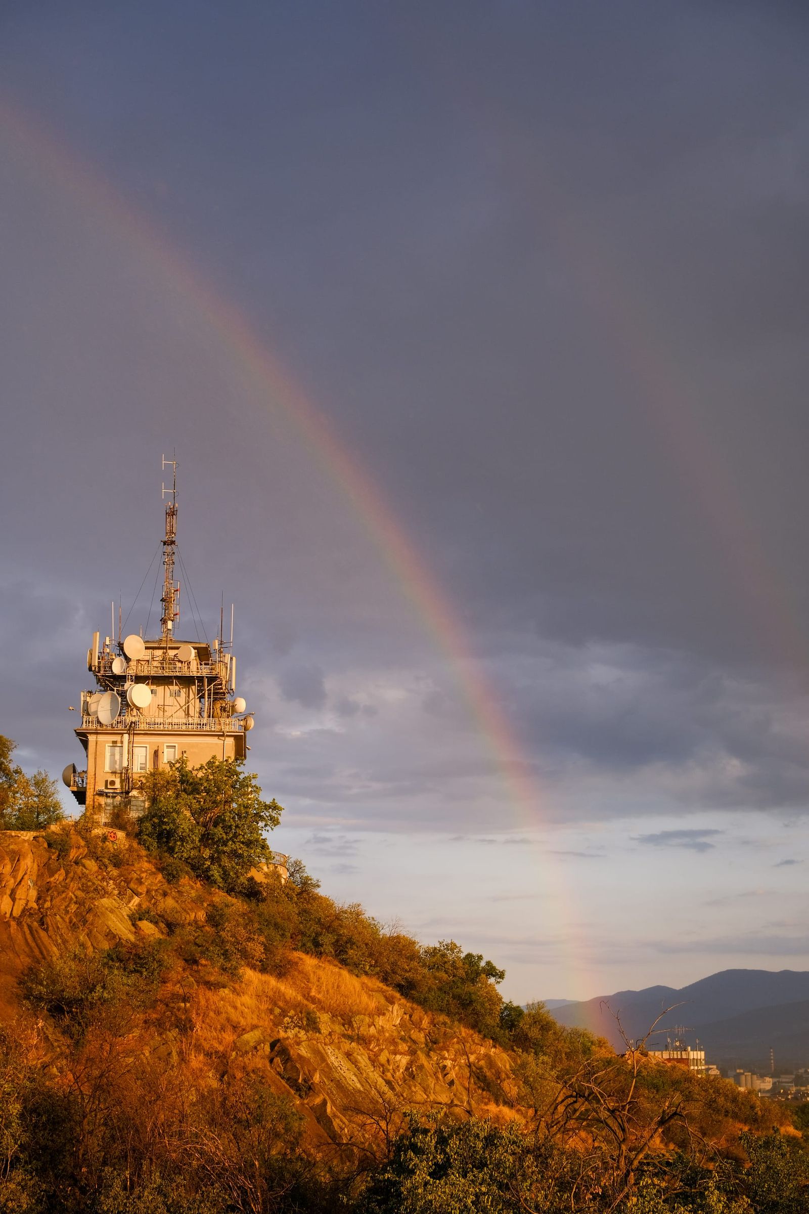 Rainbow arching over a sunlit hilltop broadcast tower beneath dark rain clouds