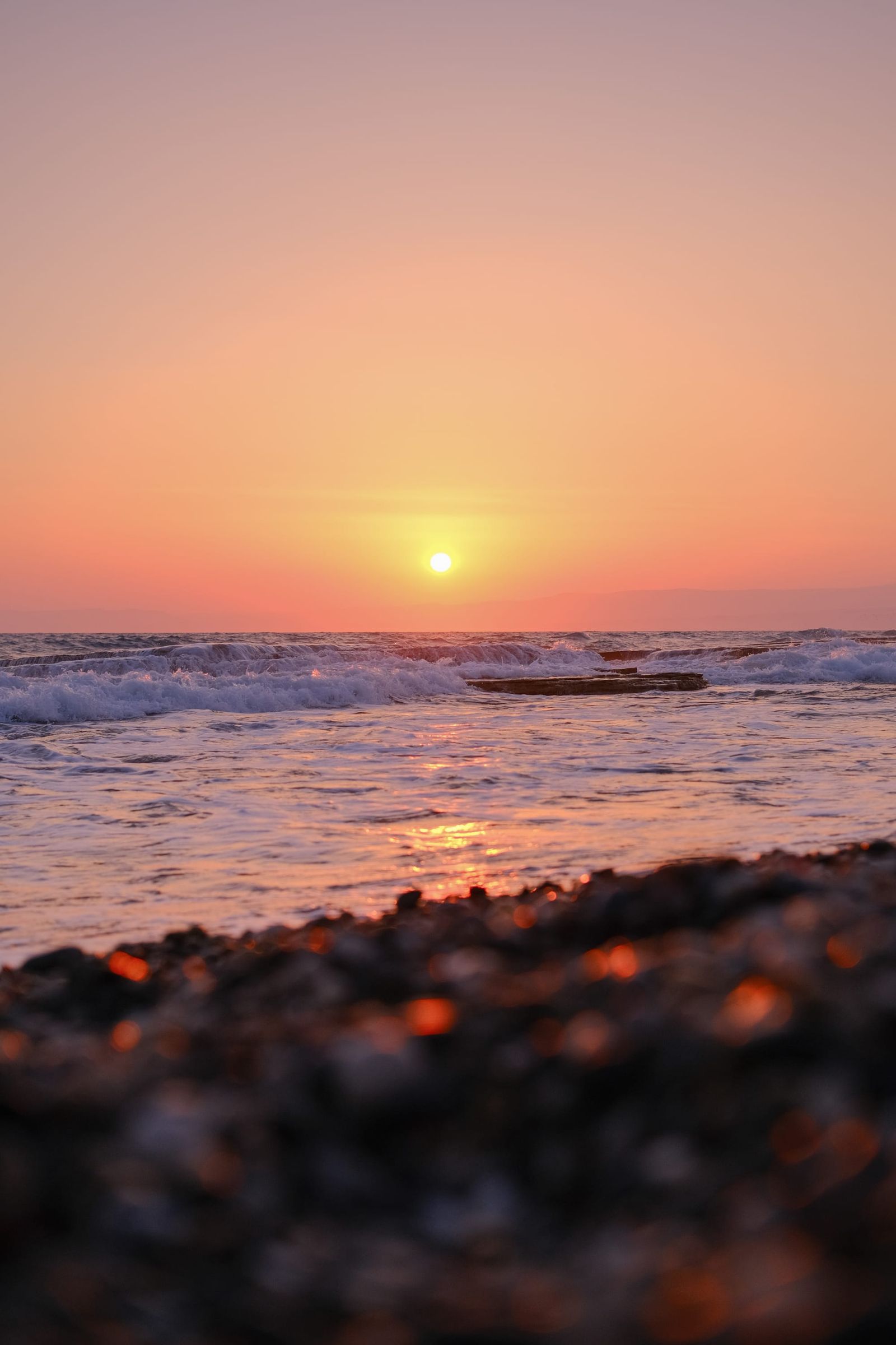 Orange sun setting over gentle sea waves and a dark pebbled shore