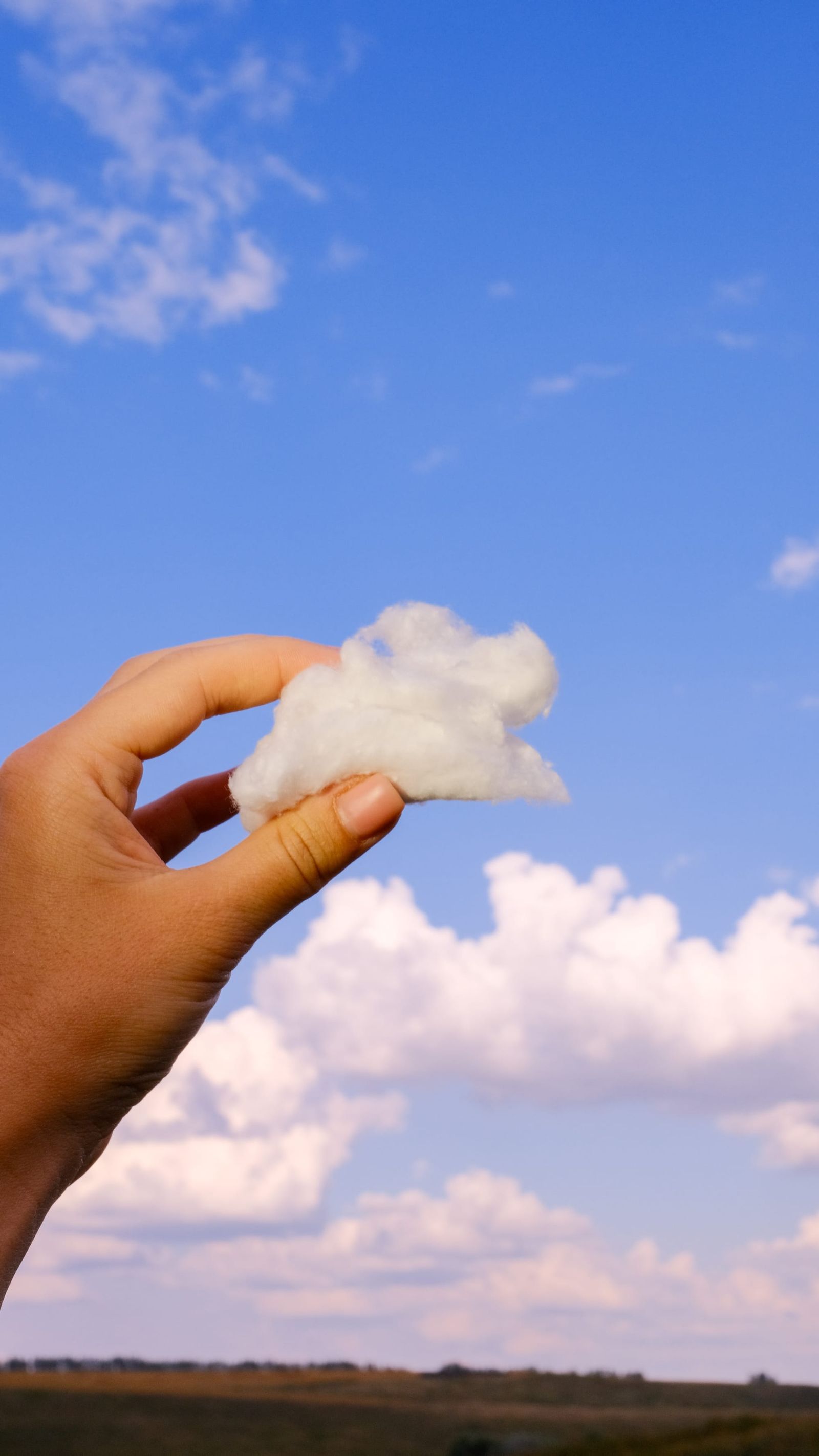 Hand holding a tuft of cotton against matching white clouds and blue sky