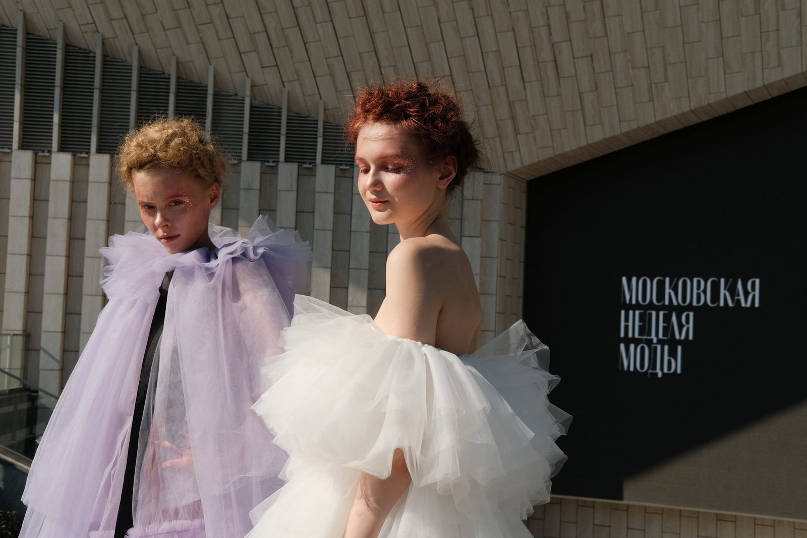 Two models pose outdoors in a lilac cape and a layered white ruffled dress.
