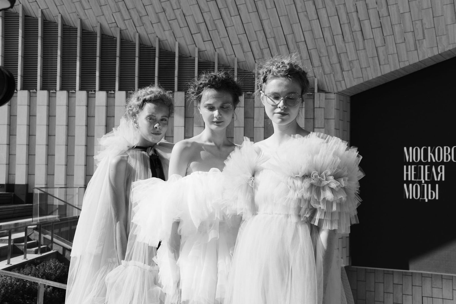 Black-and-white group portrait of three models in voluminous ruffled outfits outside the venue.