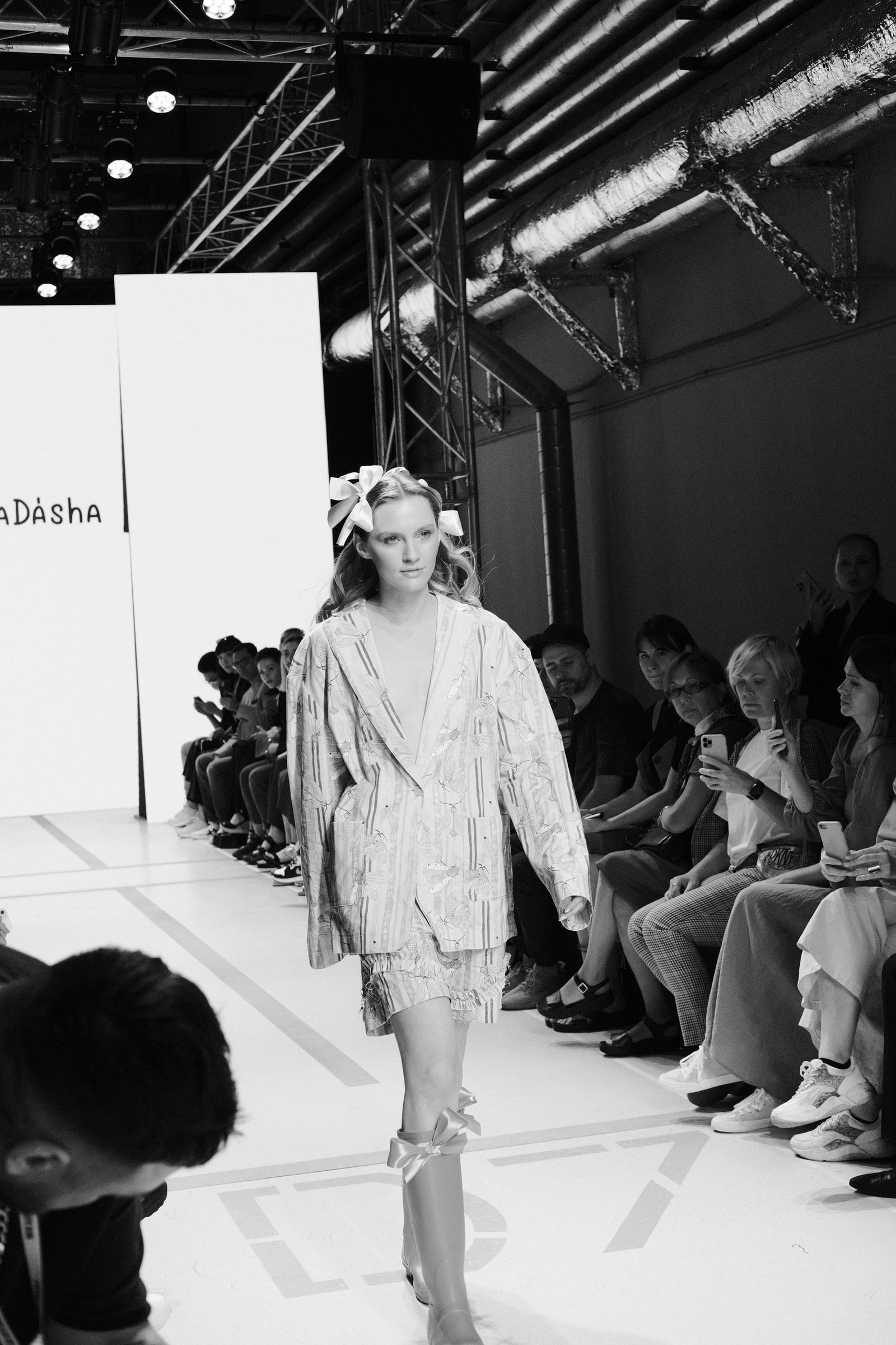 Black-and-white full-length view of a model in a loose suit and wide-brimmed hat walking the runway.