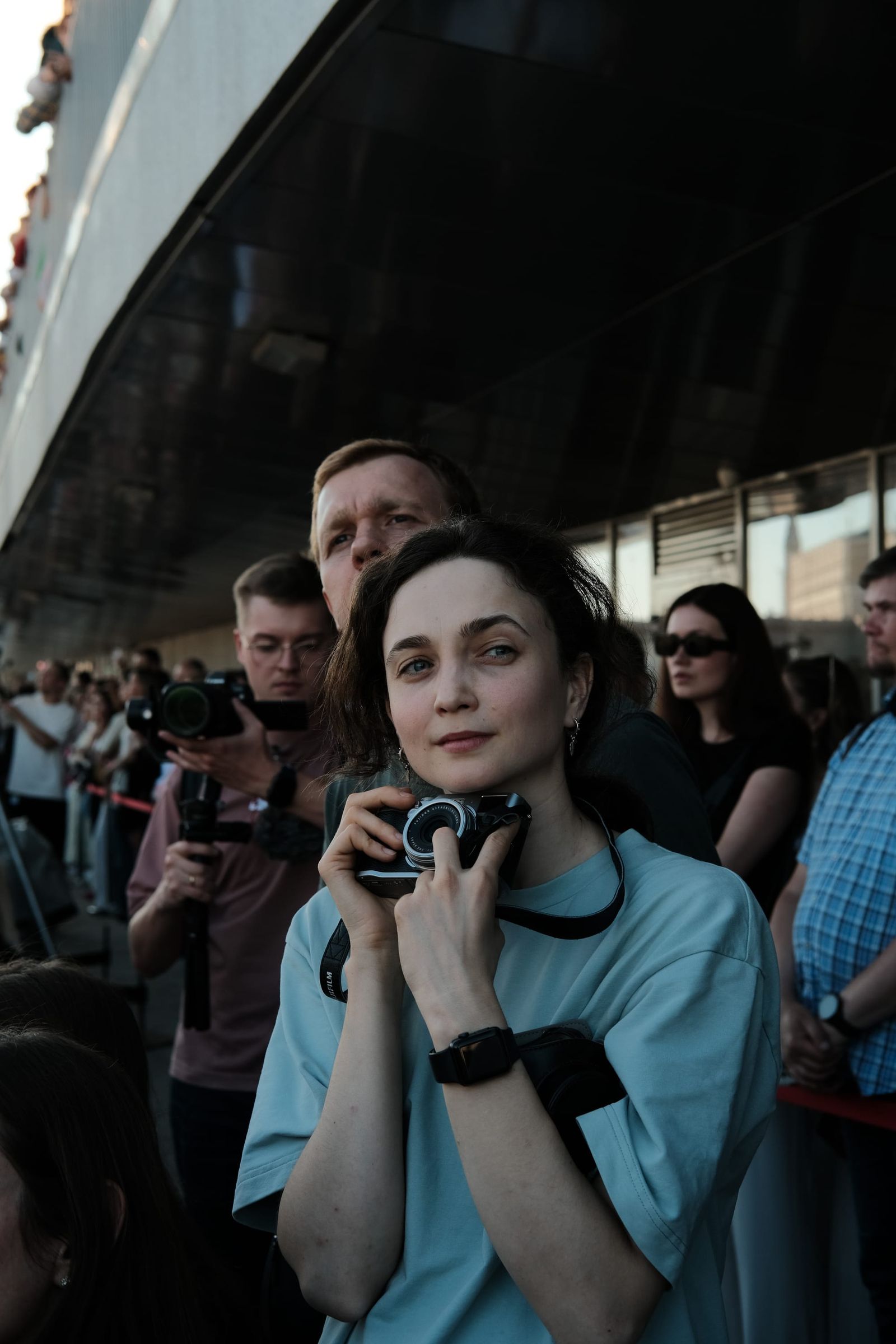 A spectator in a teal outfit sits outside with a phone in her hands, surrounded by other guests.