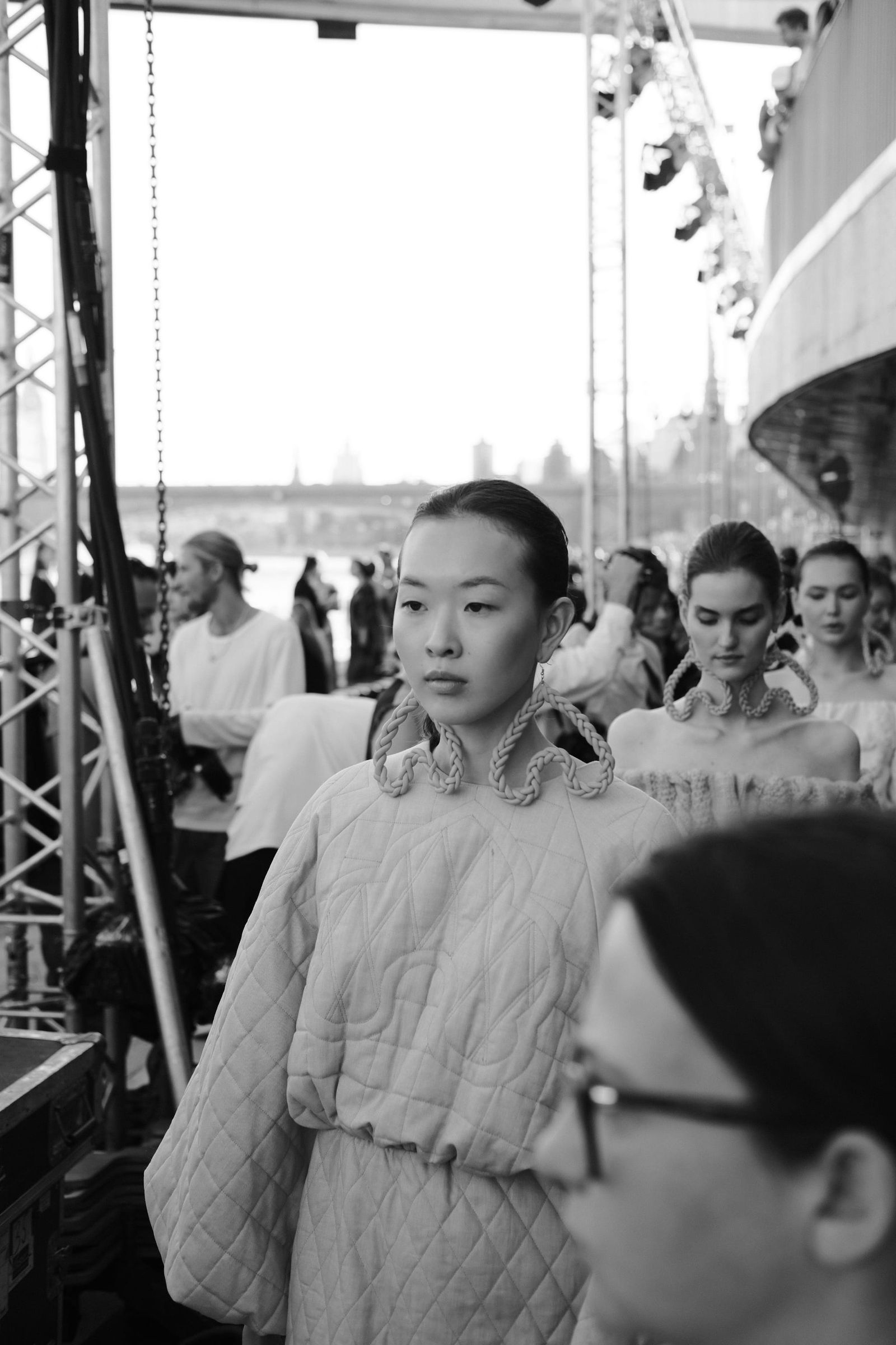 A model in a belted, textured dress passes spectators in a black-and-white photograph.