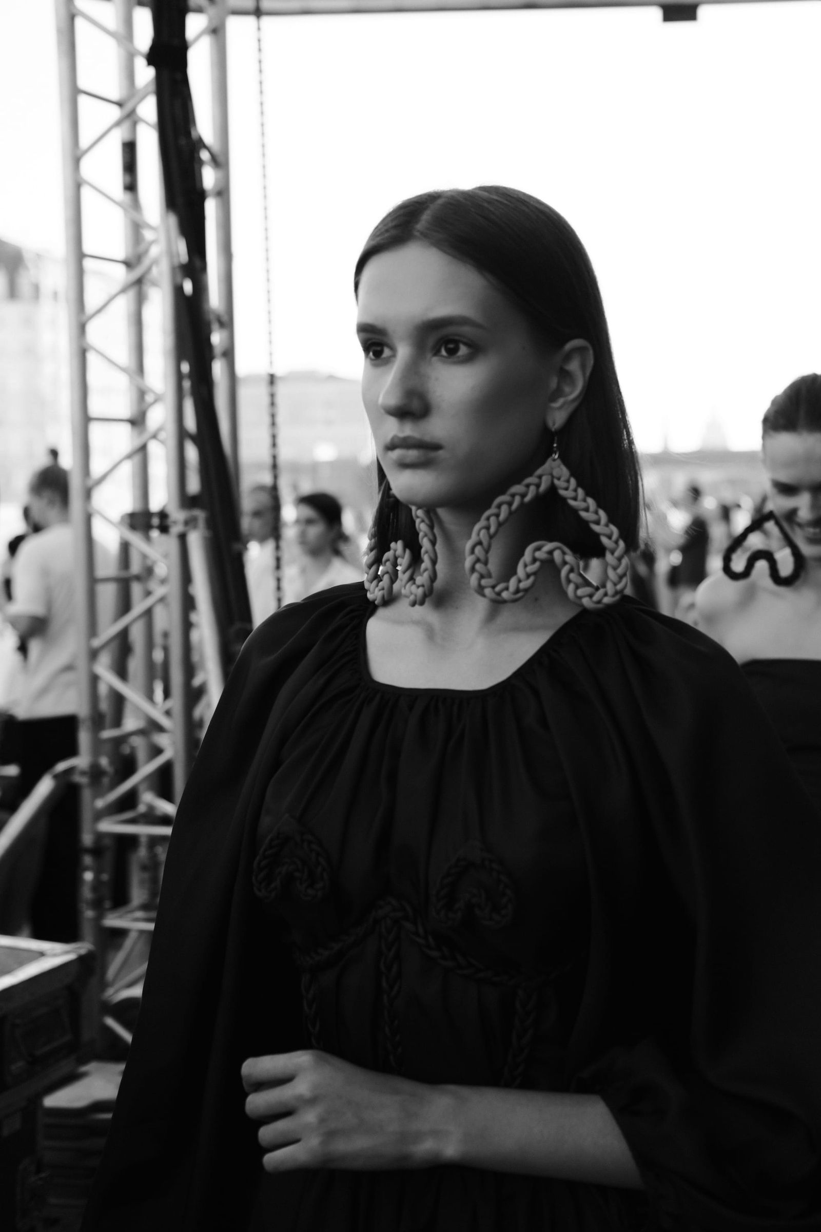 A model in a dark dress with a light ruffled collar stands near the spectators in black and white.