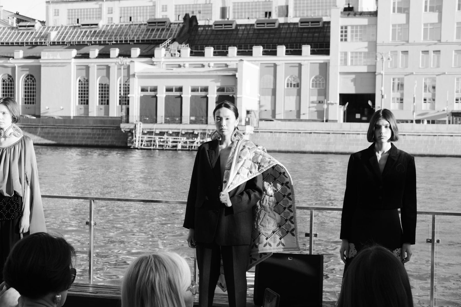 Three models stand beside the river in black and white, with waterfront buildings behind them.