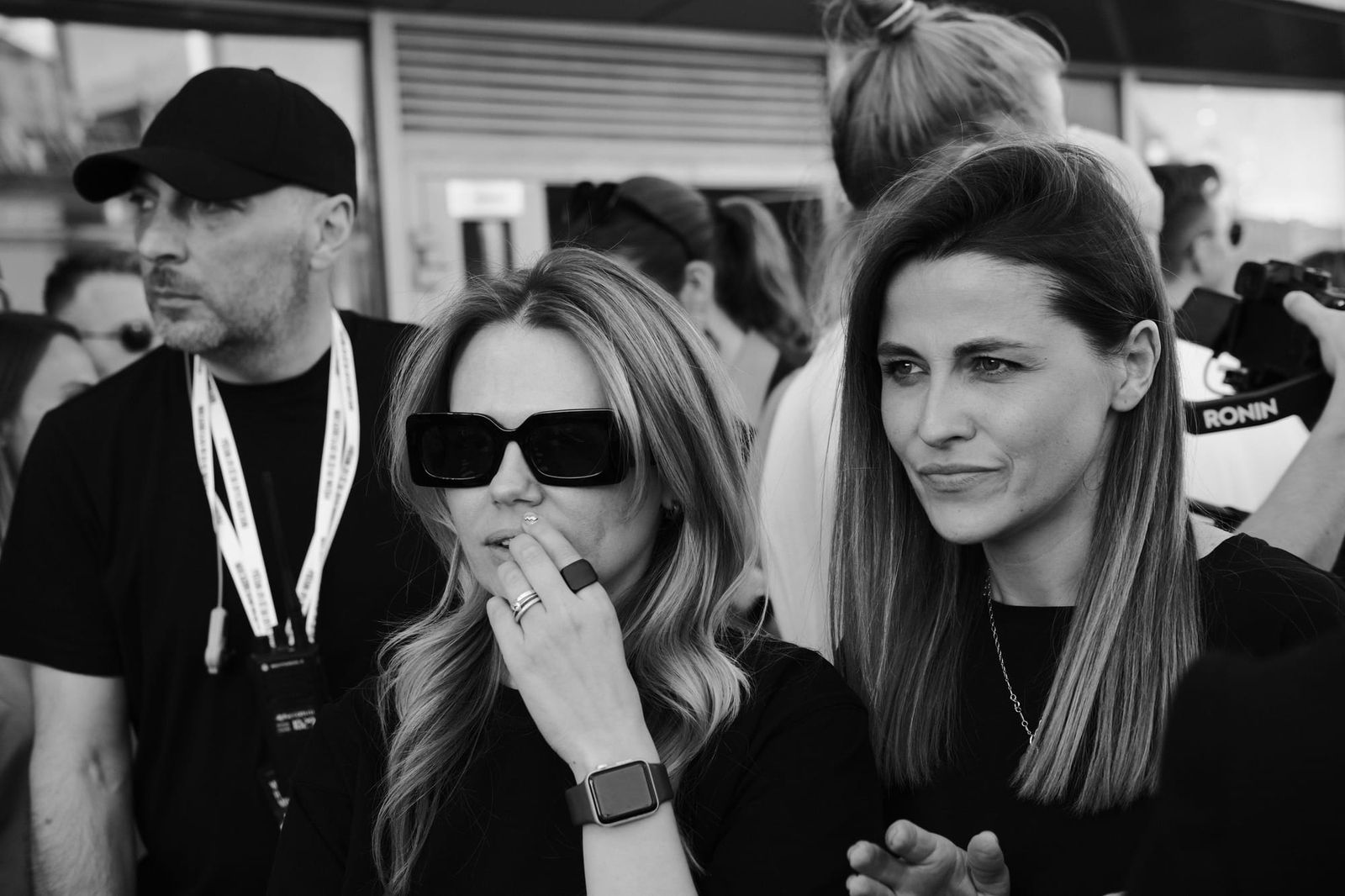 Two women stand among the crowd in black and white, one wearing dark sunglasses.