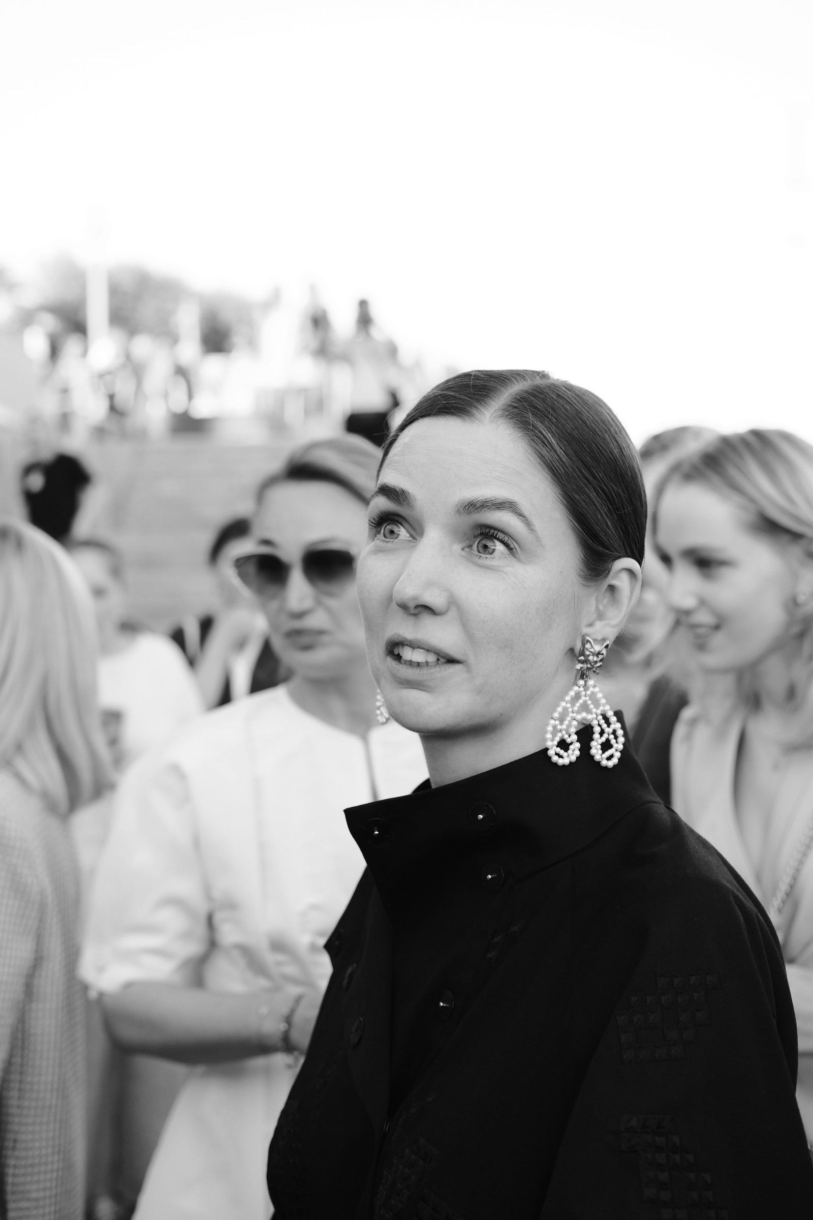 Black-and-white portrait of a woman in a high-necked dark outfit with other guests behind her.
