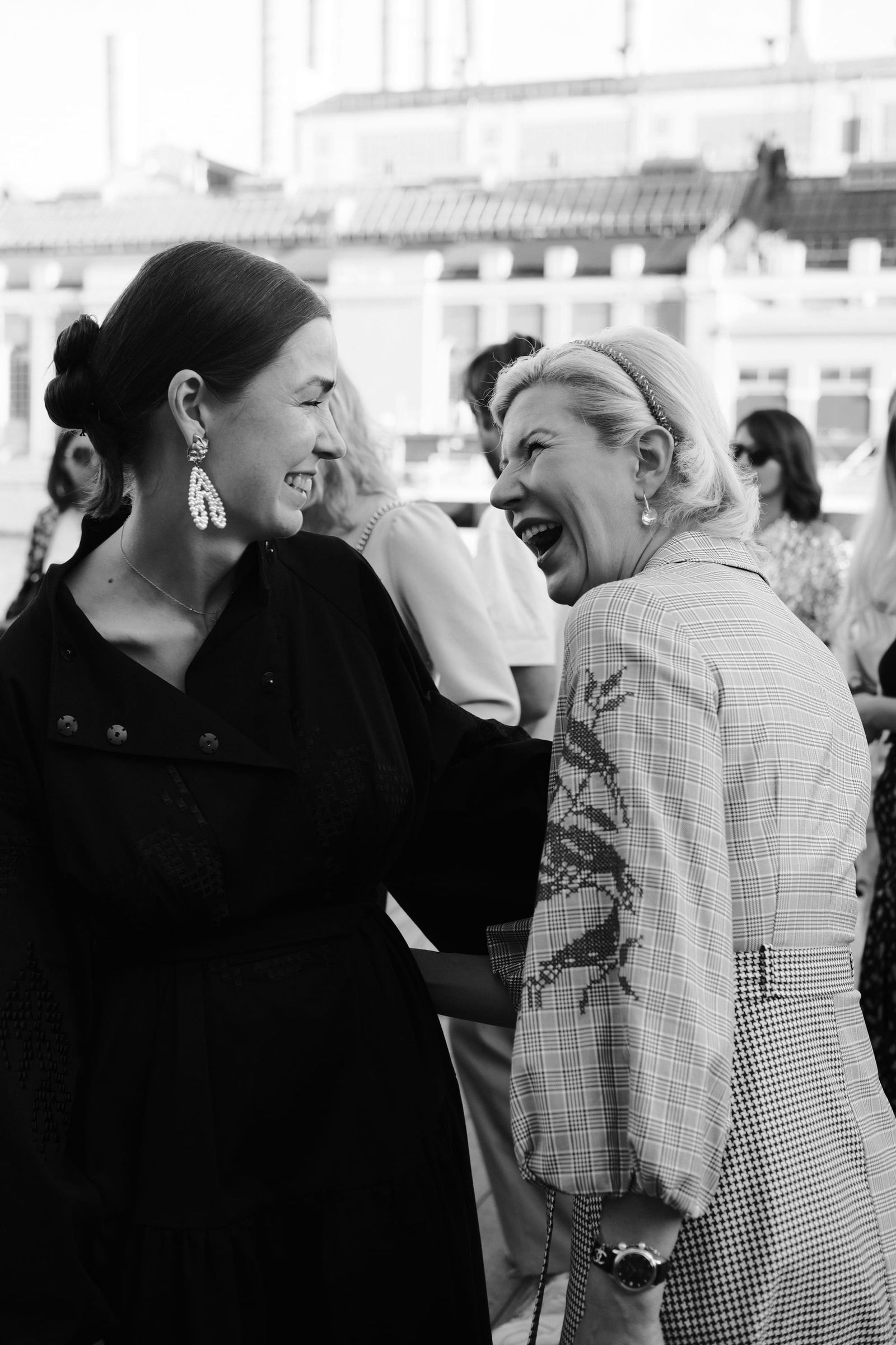 Two women talk among the guests in a black-and-white photograph, one raising a hand to her face.