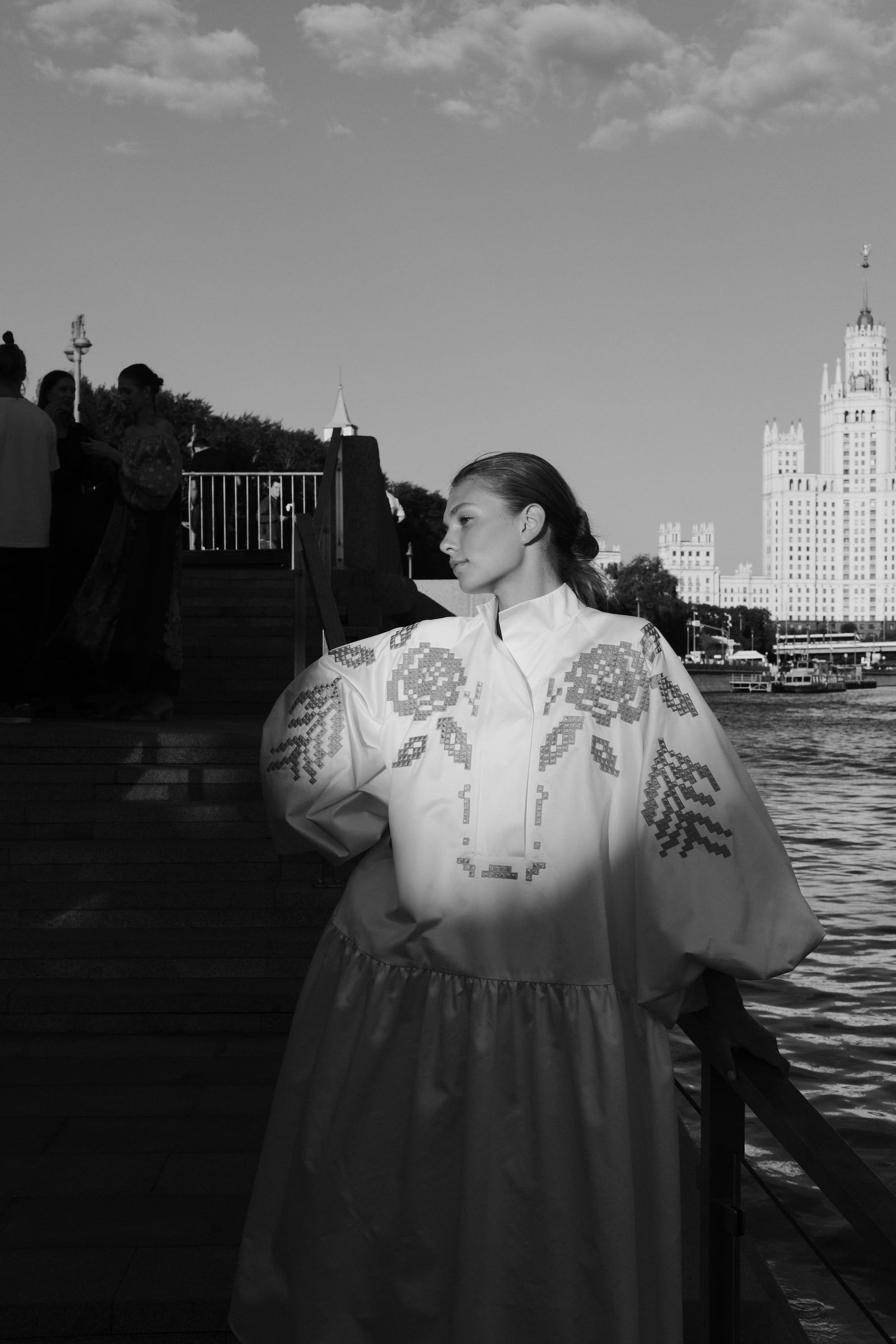 A model in a light dress with oversized flower decorations stands beside the river in black and white.