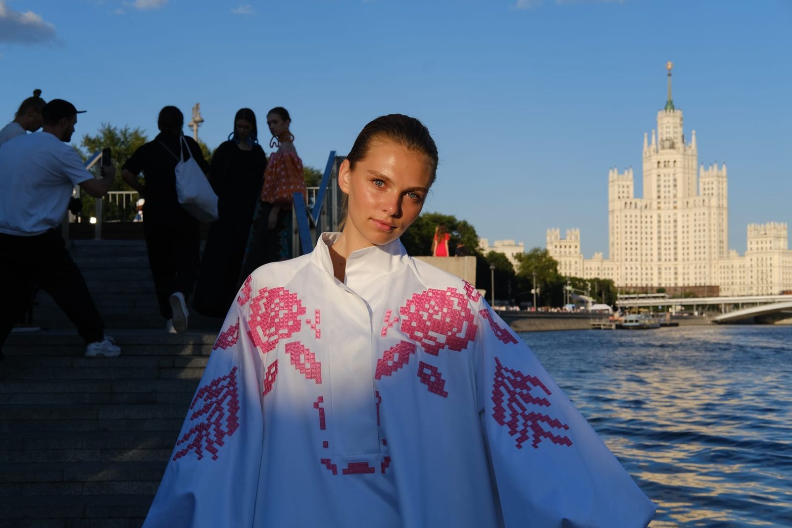 A model wears a white cape-like outfit decorated with pink flowers beside the river, with a tall building across the water.
