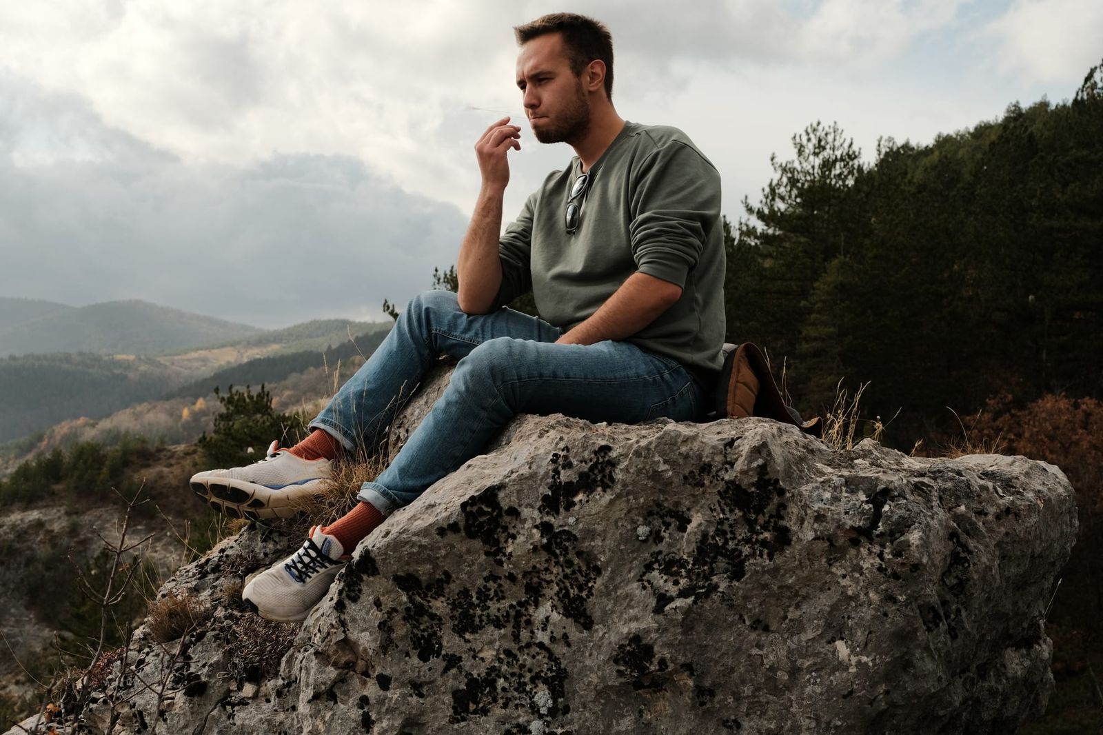 A man sits on a large rock with mountain scenery and clouded sky behind him.