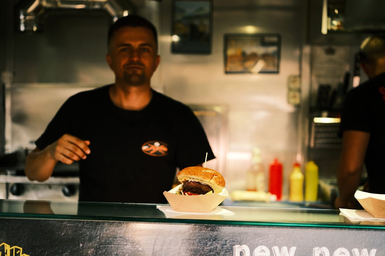 A cook stands behind a food-stall counter with a freshly prepared burger.