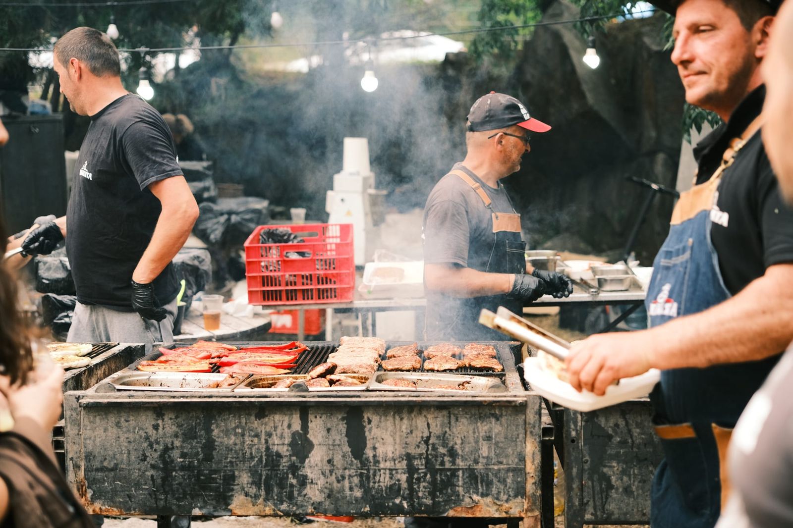 Cooks tend a wide outdoor grill as smoke rises around the food stall.