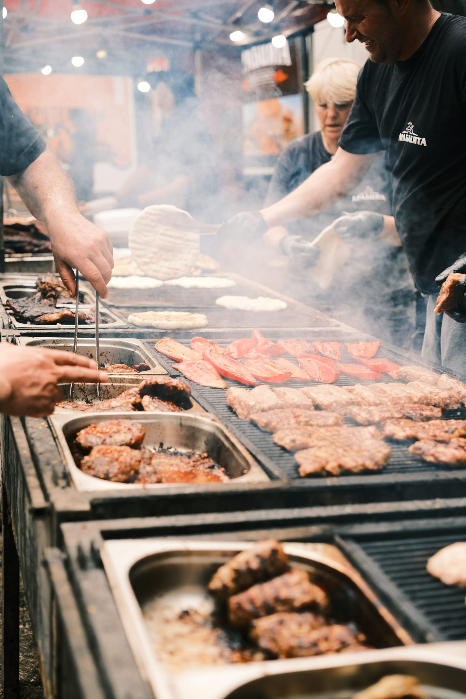 Rows of meat cook on a smoky grill beside trays of prepared food.