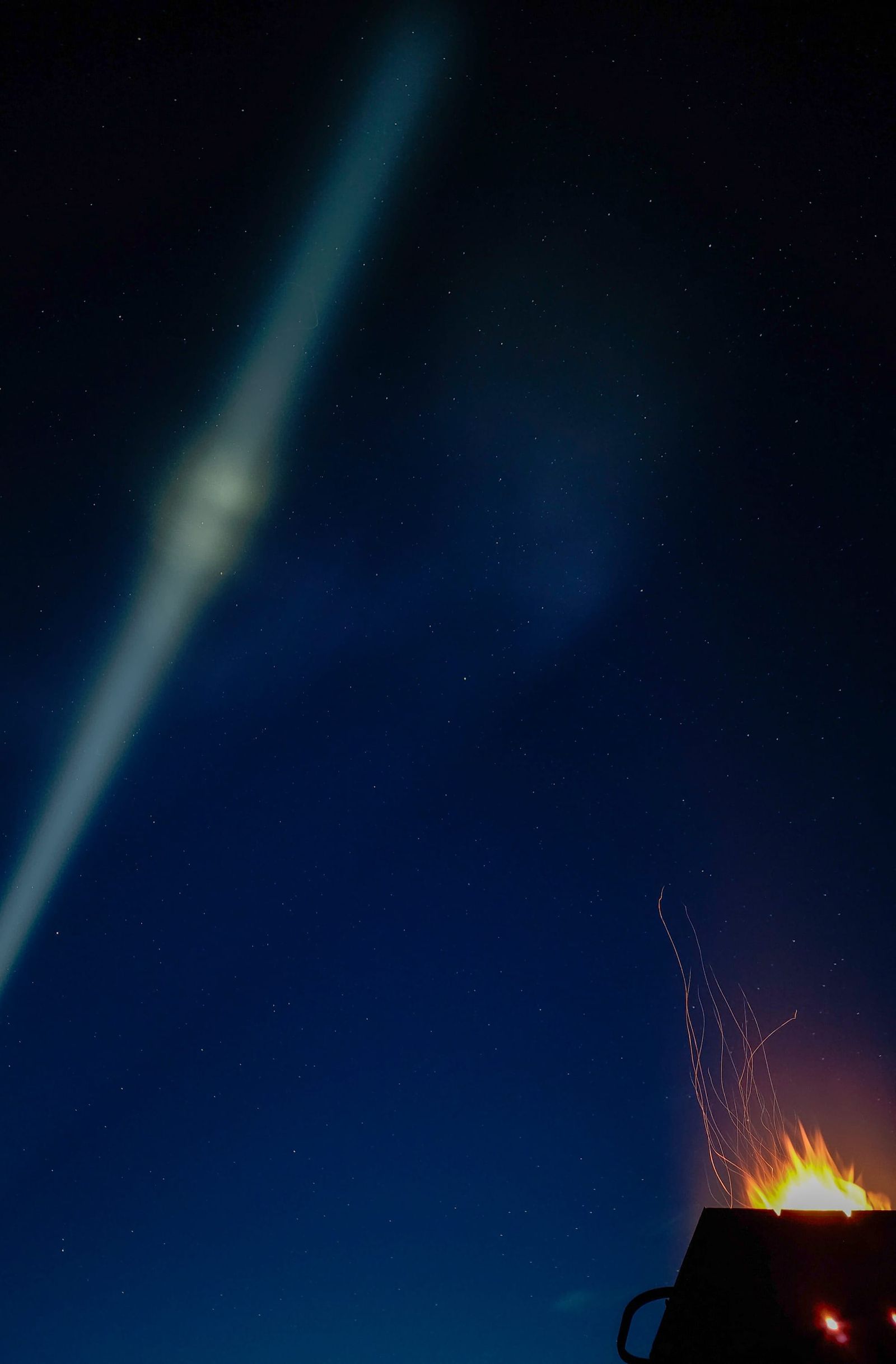 Blue-green beam crossing a dark night sky above sparks from a fire