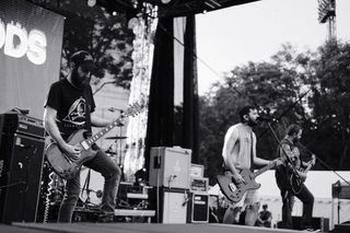 Three members of 1000Mods playing guitars and bass on an outdoor festival stage.