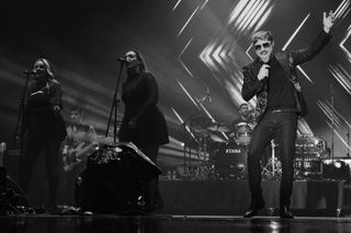 Miro singing with one arm raised beside two backing vocalists and a band onstage.