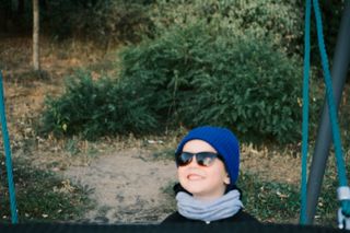 Niño sonriente con gorro azul y gafas de sol montado en un columpio al aire libre.