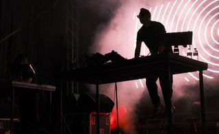 Silhouetted electronic musician at a table of equipment against pink smoke and circular stage graphics.