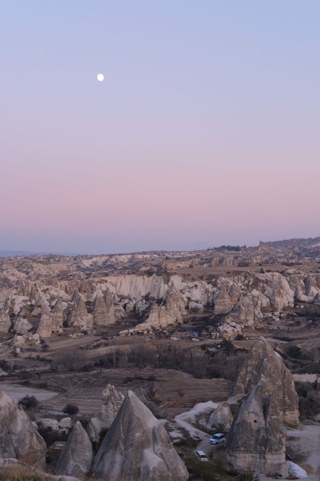 Moon above Cappadocia rock formations beneath a pale pink and blue twilight sky