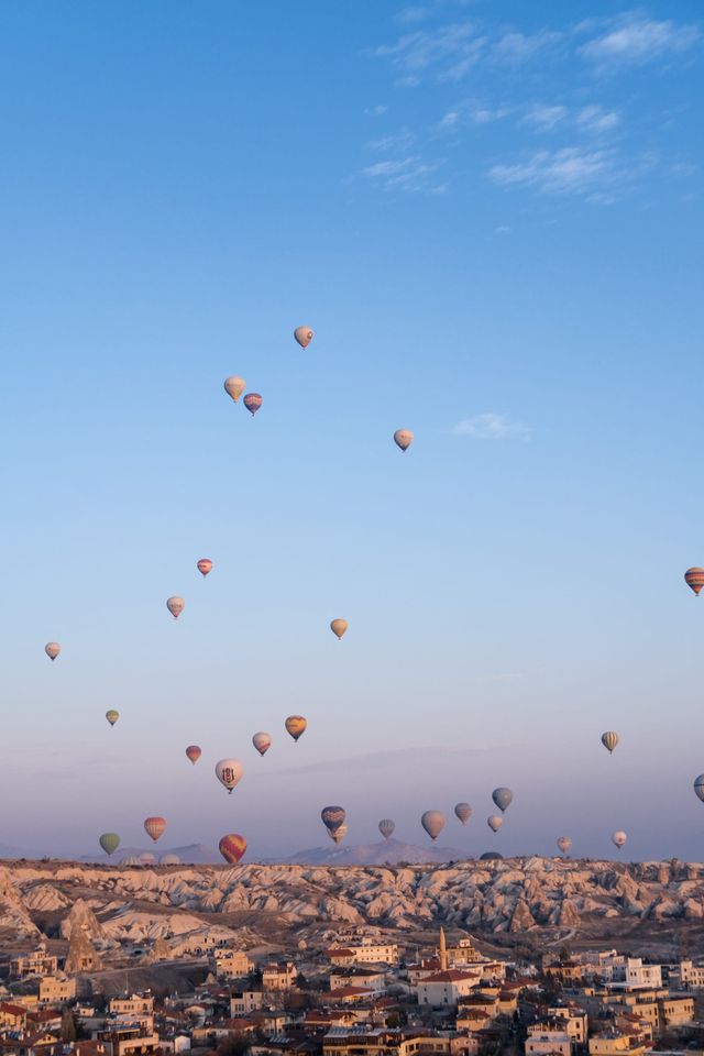 Dozens of hot-air balloons rising over a rocky town beneath a clear morning sky