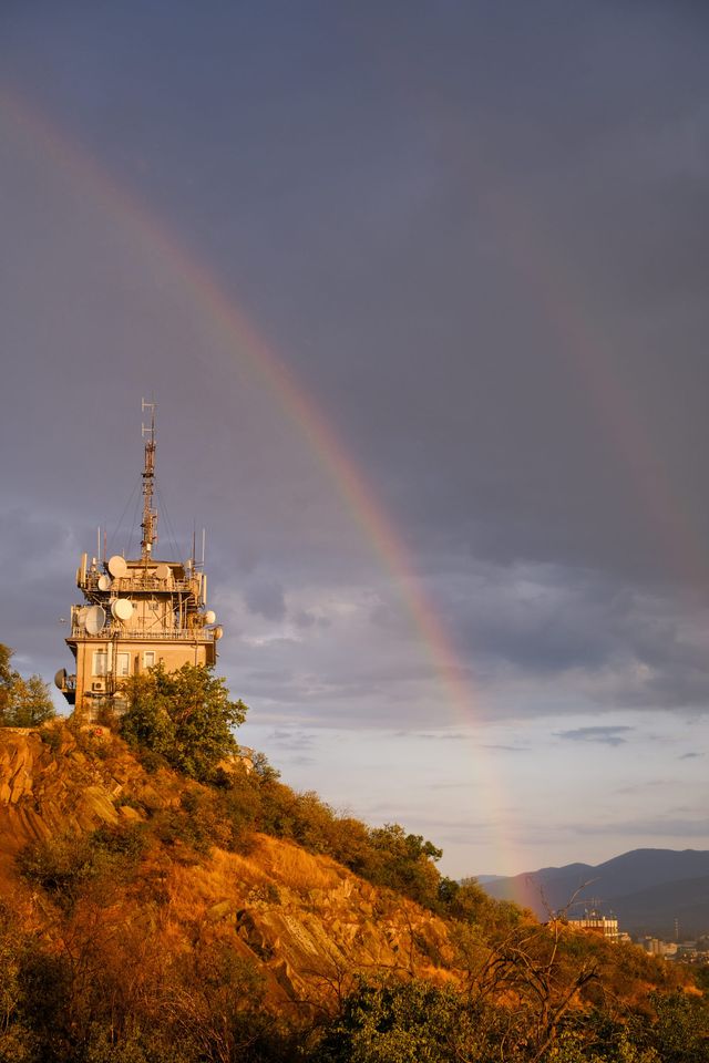 Rainbow arching over a sunlit hilltop broadcast tower beneath dark rain clouds