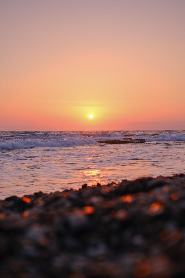 Orange sun setting over gentle sea waves and a dark pebbled shore