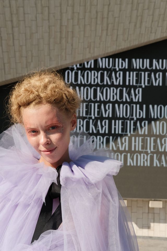 A model in a pale lilac cape stands outdoors beside the fashion-week entrance.