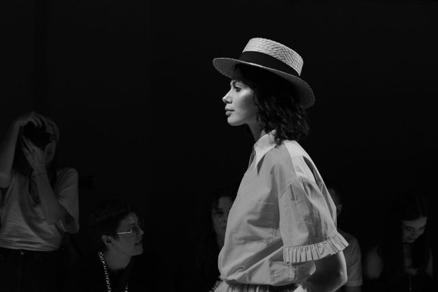 Black-and-white side view of a model in a light blouse with ruffled sleeve cuffs and a brimmed hat.