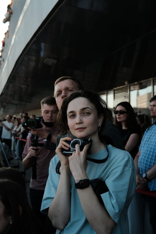 A spectator in a teal outfit sits outside with a phone in her hands, surrounded by other guests.
