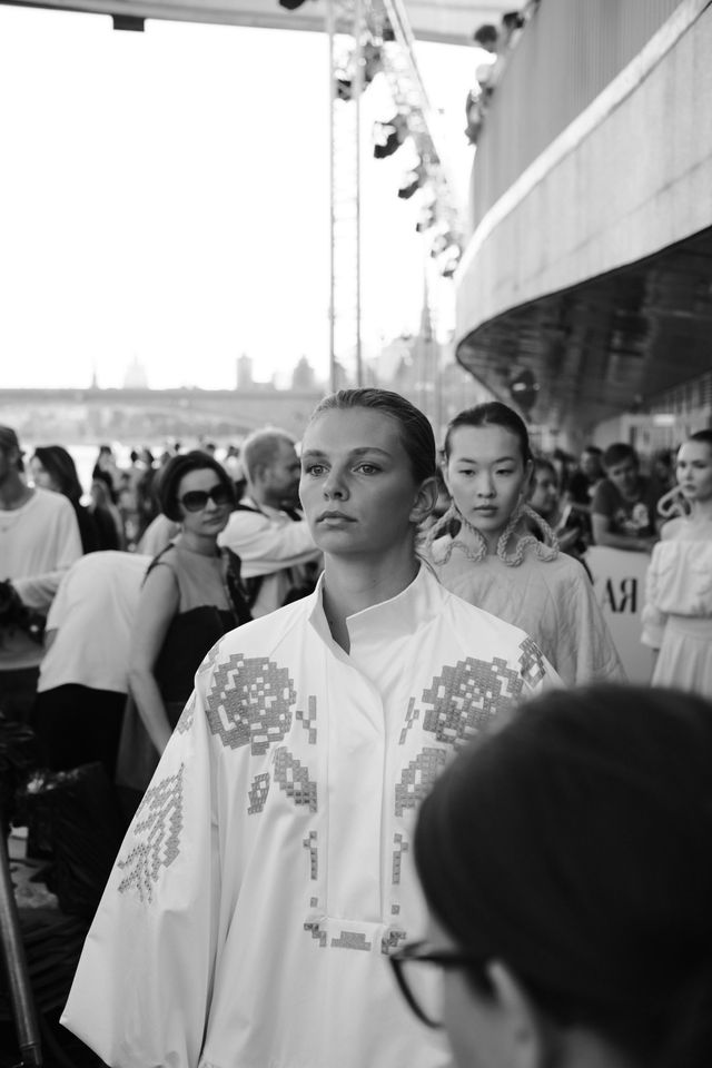 Black-and-white photograph of a model in a light dress with decorative flower-like appliqués walking through a crowd.