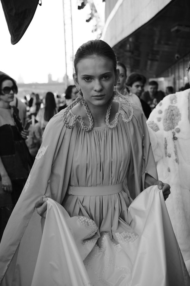 Black-and-white photograph of a model holding the sides of a long pleated dress.