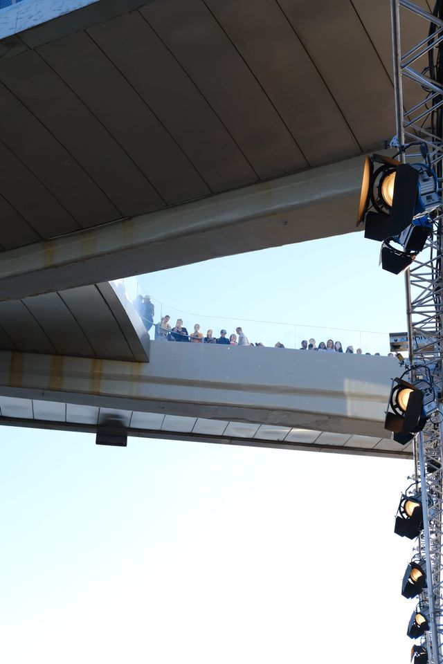 Looking upward at the underside of an elevated walkway and suspended event lights beneath a blue sky.