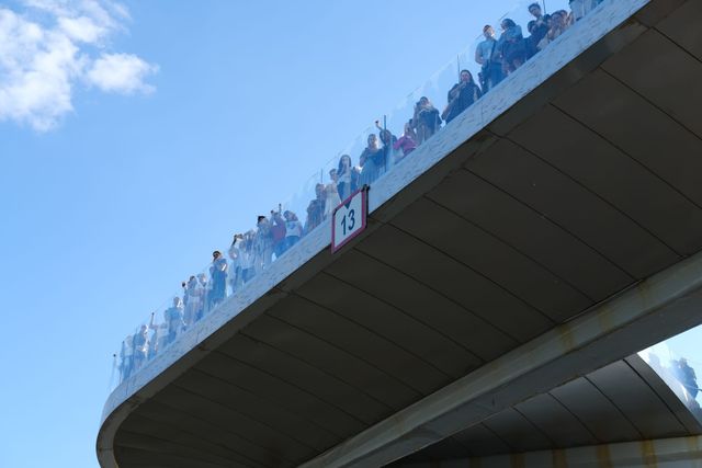 Spectators line the glass edge of an elevated walkway high above the event, against blue sky and white clouds.