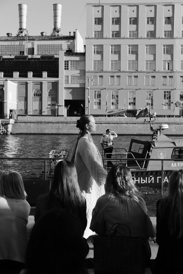 Black-and-white photograph of models presenting their outfits beside the river, with spectators in the foreground.