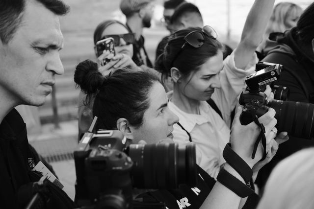 Cameras and photographers surround two women in a tightly framed black-and-white image.