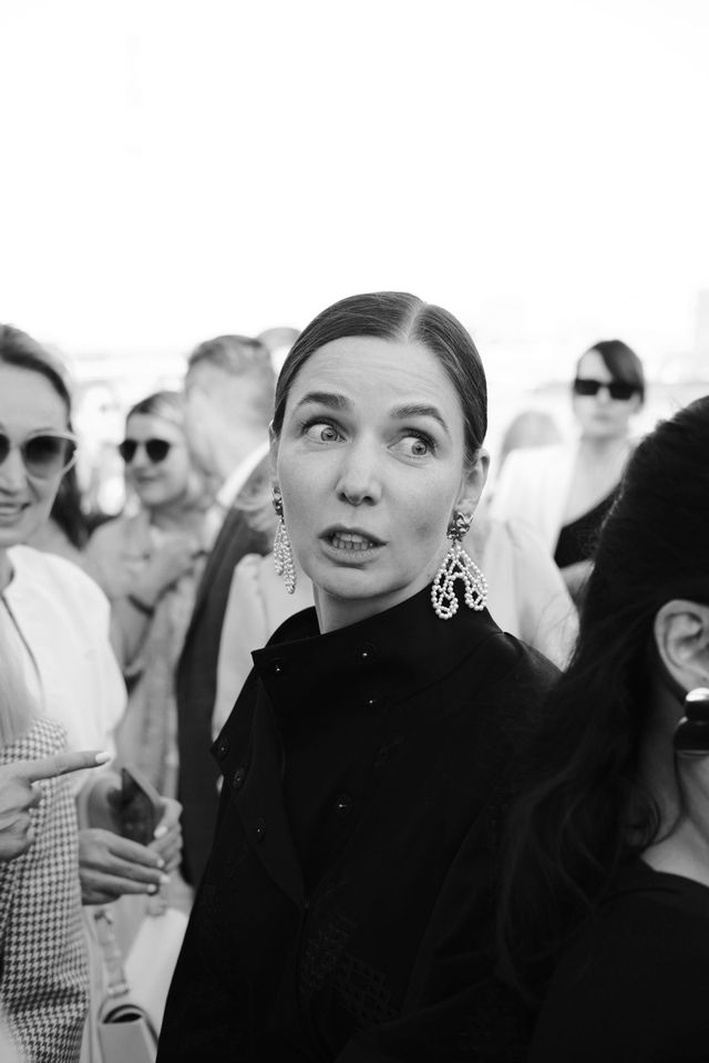A woman in a high-necked dark outfit looks upward while standing among the crowd in black and white.