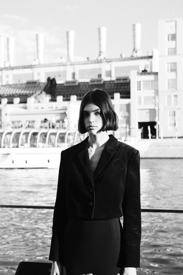 Black-and-white portrait of a model in a tailored dark suit standing at the riverside railing.