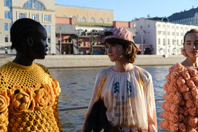 Three models wear brightly coloured knitwear and appliquéd outfits beside the river in warm sunlight.