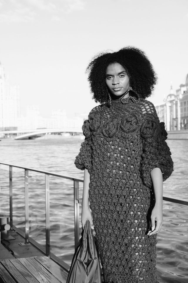 Black-and-white full-length portrait of a model in a long, textured dress beside the river.