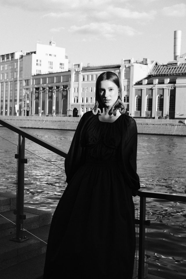 A model in a long dark dress with a ruffled neckline stands at a riverside railing in black and white.