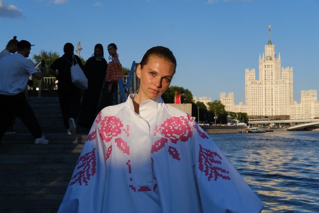 A model wears a white cape-like outfit decorated with pink flowers beside the river, with a tall building across the water.