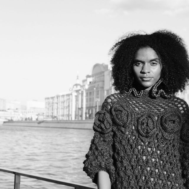 Black-and-white square portrait of a model with curly hair wearing a richly textured dark outfit by the river.